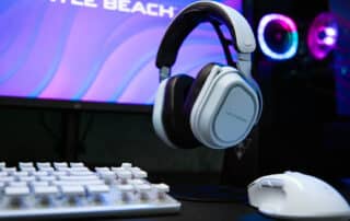 Gaming-opsætning med en computerskærm med lilla grafik, et hvidt tastatur, en hvid gaming-mus og Turtle Beach-headset på et stativ.