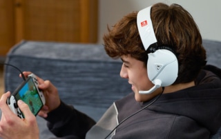 En person med et hvidt headset sidder i en sofa og er fuldt fokuseret på sin håndholdte spillekonsol, mens en automatisk klædefunktion gemmer deres spilfremskridt i baggrunden.
