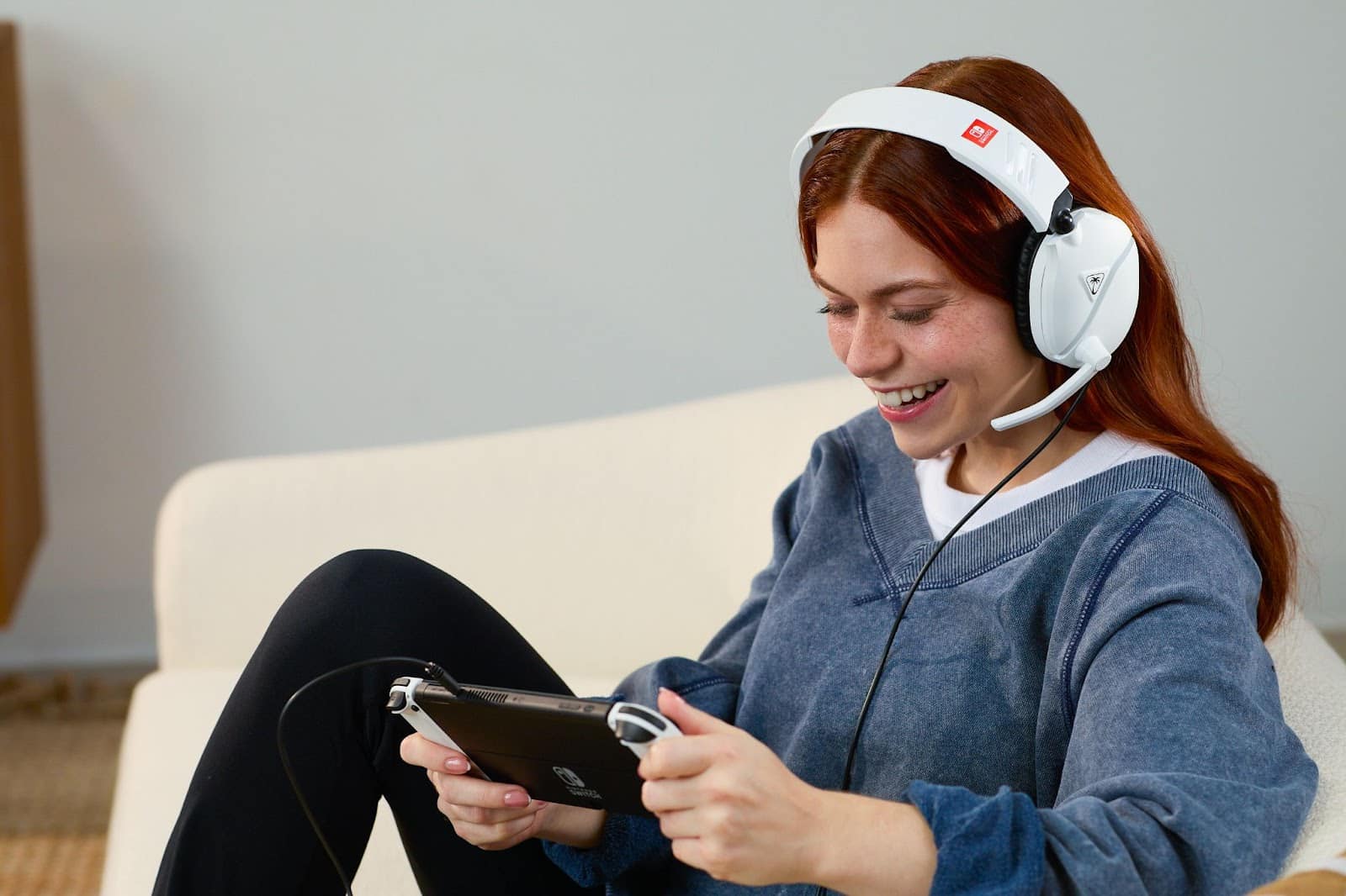 En person med et hvidt headset smiler, mens han holder og spiller på en håndholdt spilleenhed, siddende på en lys sofa. En person med et hvidt headset smiler, mens han holder og spiller på en håndholdt spilleenhed, siddende på en lys sofa.