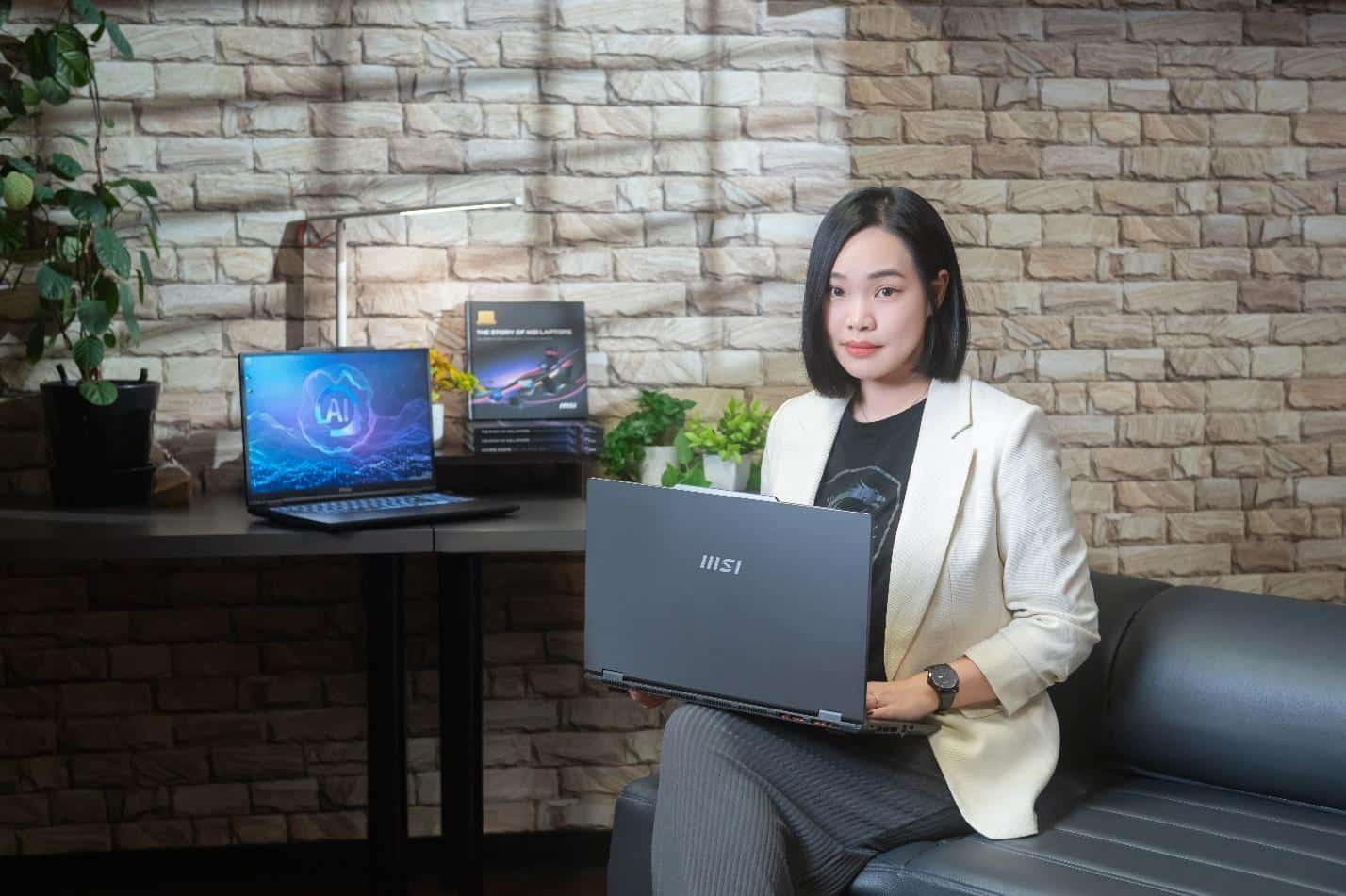 En kvinde sidder i en sofa med en MSI-laptop i hånden, mens en anden VenturePro-laptop og en reklameskærm står på et bord ved siden af hende op ad en stenbelagt væg. En kvinde sidder i en sofa med en MSI-laptop i hånden, mens en anden VenturePro-laptop og en reklameskærm står på et bord ved siden af hende op ad en stenbelagt væg.
